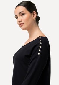 Top nero a maniche lunghe con scollatura a V, caratterizzato da bottoni color crema lungo la cucitura della spalla. Tessuto liscio con una silhouette aderente.