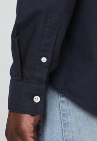 Granatowa koszula z długim rękawem, zapinana na białe guziki, o gładkiej fakturze. Mankiety mają jedno guziki zapięcie.