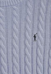 Jasnoniebieski sweter z dzianiny warkoczowej z wypukłymi skręconymi wzorami i małym czarnym haftem logo na przodzie.