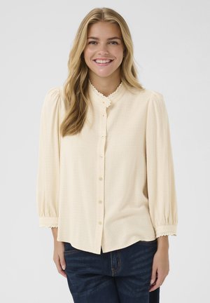 Hellbeige Bluse mit Knöpfen, die ein strukturiertes Karomuster aufweist, einen mit Spitze verzierten Kragen und Puffärmel hat. Trägt sie mit dunklen Jeans.