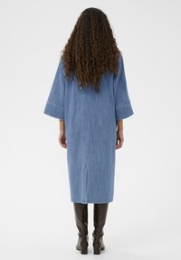 Denim obleka v svetlo modri barvi, srednje dolžine z širšimi rokavi in razporkom na sredini hrbta. Združena z rjavimi gumijastimi škornji do kolen z vzorcem.