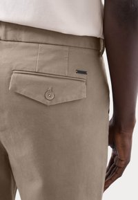 Pantalones beige con una tela texturizada, que presentan un bolsillo trasero triangular con cierre de botón y una pequeña etiqueta negra que dice "AUTOGRAPH."