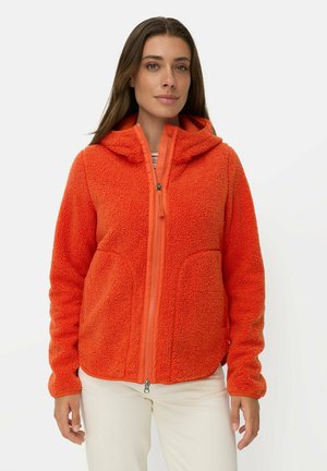 Kvinde iført en lys orange fleecejakke med hætte og hvide bukser, stående mod en ensartet lys baggrund.