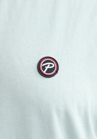 Maglietta di cotone azzurro chiaro con una toppa rotonda che presenta un logo nero, rosso e bianco. Tessuto liscio, maniche corte e collo rotondo.