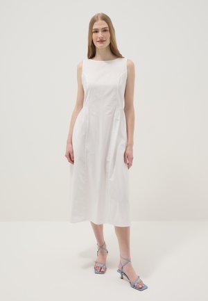 Jeune femme debout portant une robe midi blanche sans manches et des sandales à talons bleu clair avec brides, sur fond uni.