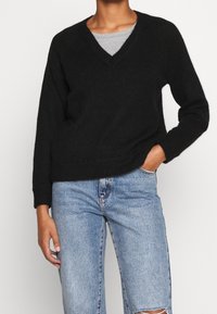 Schwarzer gestrickter Pullover mit V-Ausschnitt, langen Ärmeln und gerippten Bündchen. Über einem grauen Oberteil getragen, kombiniert mit hellblauen, verwaschenen Jeans.