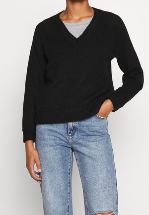 Czarny sweter z dzianiny z dekoltem w kształcie litery V, długimi rękawami i ściągaczami. Nosi się na szarym topie, połączony z jasnoniebieskimi dżinsami z efektami sprania.
