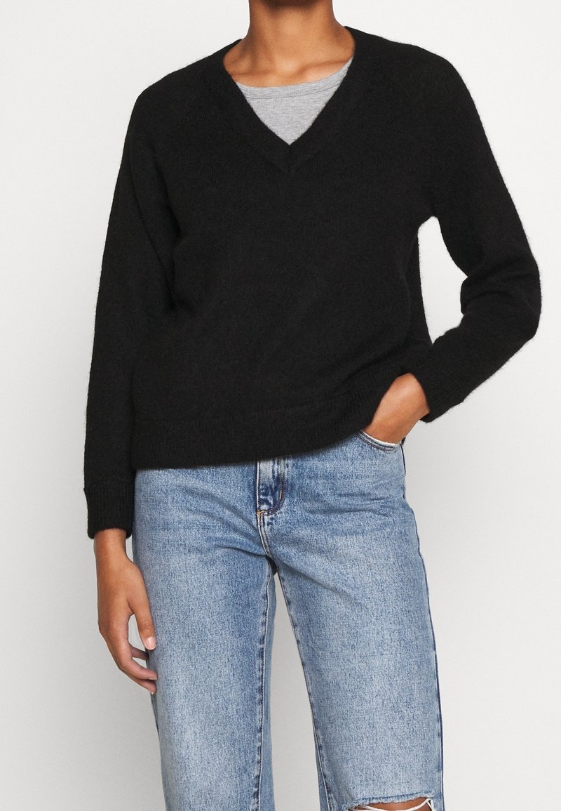 Schwarzer gestrickter Pullover mit V-Ausschnitt, langen Ärmeln und gerippten Bündchen. Über einem grauen Oberteil getragen, kombiniert mit hellblauen, verwaschenen Jeans.