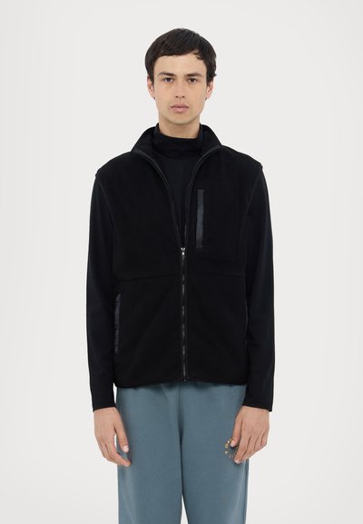 Gilet en polaire noir avec col haut, fermeture éclair sur le devant, deux poches latérales et une poche sur la poitrine, doté d'une texture lisse et d'un design minimaliste.