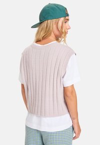 Gilet en tricot rose avec une texture côtelée verticale, un col rond et une conception sans manches, superposé sur une chemise blanche et un pantalon à carreaux.
