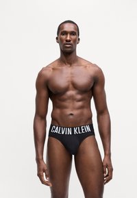 Crne pamučne gaćice s elastičnom trakicom koja ističe natpis "CALVIN KLEIN" u bijeloj boji. Model stoji u neutralnoj pozi protiv jednostavne pozadine.