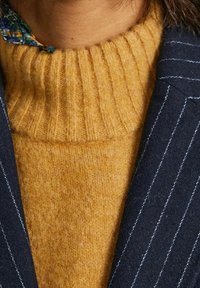Close-up of a person wearing a mustard yellow ribbed turtleneck sweater and a navy blue pinstripe blazer.