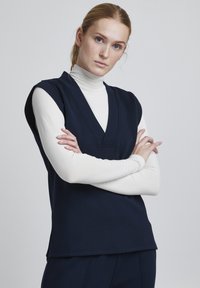Top senza maniche di colore blu navy con scollo a V, sovrapposto a un dolcevita bianco. Tessuto morbido, vestibilità comoda e orlo dritto con spacchi laterali.