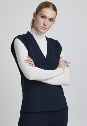 Top senza maniche di colore blu navy con scollo a V, sovrapposto a un dolcevita bianco. Tessuto morbido, vestibilità comoda e orlo dritto con spacchi laterali.
