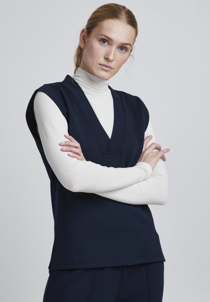 Top senza maniche di colore blu navy con scollo a V, sovrapposto a un dolcevita bianco. Tessuto morbido, vestibilità comoda e orlo dritto con spacchi laterali.