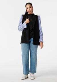 Zwarte mouwloze op maat gemaakte vest over een zwart kant top en een blauw gestreepte blouse met pofmouwen. Lichtblauwe wijde jeans en witte sneakers.