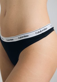 Črne tangice iz mehkega blaga, s belim elastičnim pasom z blagovno znamko "Calvin Klein" v črni barvi. Gladka tekstura, nizko krojena.