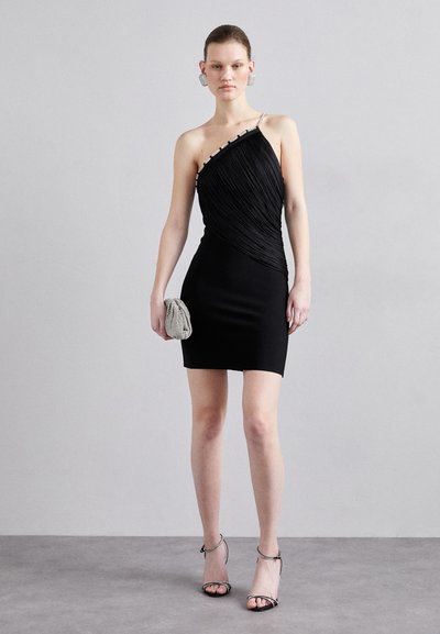 Robe mini noire avec un design plissé asymétrique, une bretelle unique et des accents argentés, accompagnée d'une petite pochette métallique et de talons à lanières.