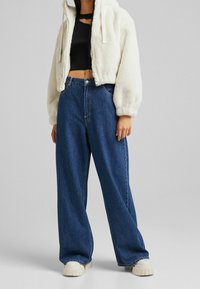 Luddig vit kort jacka med huva, tillsammans med högmidjade blå vida jeans och klumpiga vita sneakers. Slät tygtextur.