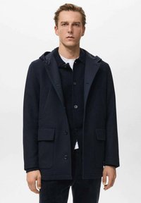 Joven vestido con un abrigo con capucha azul marino sobre un cárdigan negro abotonado y una camisa blanca, de pie contra un fondo blanco liso.