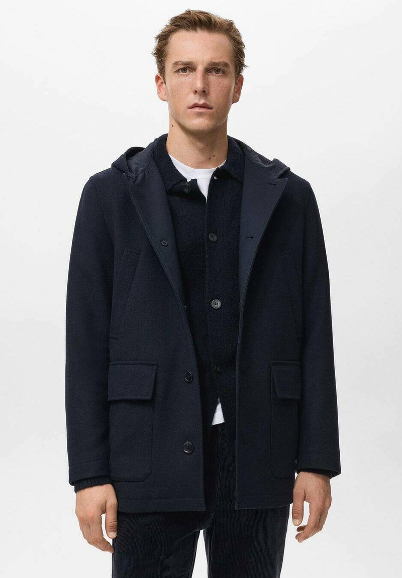 Joven vestido con un abrigo con capucha azul marino sobre un cárdigan negro abotonado y una camisa blanca, de pie contra un fondo blanco liso.
