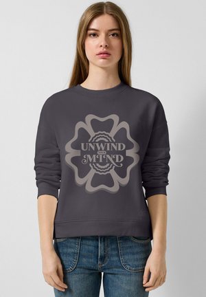 Il pullover grigio presenta un grande grafico centrale con il testo "DISCONNETTI LA TUA MENTE", circondato da una forma floreale. Tessuto morbido.