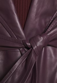 Nahaufnahme eines gebundenen dunkelpurpurnen Ledergürtels über einem passenden Lederledermantel mit einem dunkelburgunderfarbenen gerippten Pullover darunter.