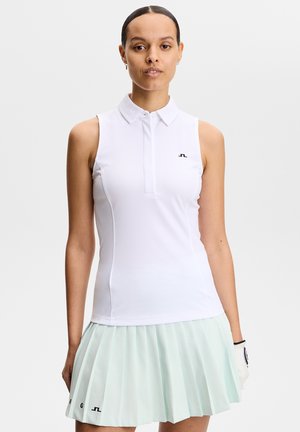 Vrouw die een mouwloze witte poloshirt en een licht mintgroene geplooide rok draagt, staand met een golfhandschoen aan haar linkerhand.