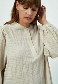 Blusa color crema in tessuto testurizzato, con colletto alto, chiusura a bottoni e maniche a sbuffo. Design a pieghe sottili in tutto il capo.