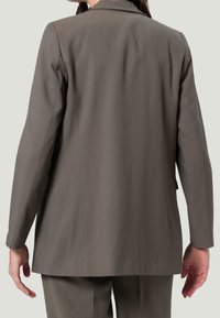 Blazer cintré gris à manches longues, avec une fente au dos unique et des lignes épurées. Fabriqué en tissu lisse, conçu pour une silhouette ajustée.