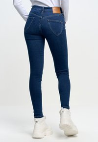 Jeans ajustados de denim azul oscuro con una cintura alta, que cuentan con dos bolsillos traseros y una etiqueta de cuero marrón en la parte posterior. Combinados con zapatos blancos.