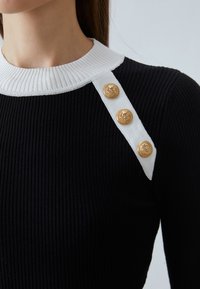 Černý žebrovaný svetr s bílým límcem a bílým ramenním popruhem se třemi zlatými knoflíky, který má strukturovaný design.