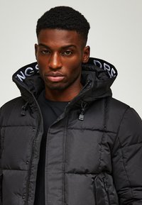 Schwarze Daunenjacke mit durchgehendem Reißverschluss, verstellbare Kapuze und strukturiertem Stoff. Verfügt über seitliche Taschen und kontrastierende Details am inneren Kragen.