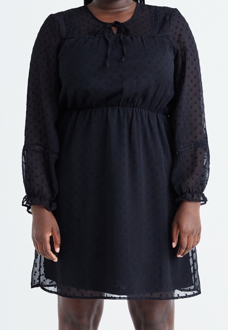 Robe noire mi-longue avec manches longues transparentes et texture à pois, avec un col noué et une taille froncée.