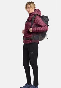 Giacca a vento color maroon, zaino nero, pantaloni neri e scarpe blu; persona in piedi di lato con la mano posizionata sul fianco, mostrando l'abbigliamento.