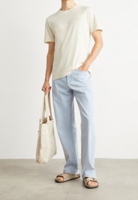 Camiseta de algodón beige, jeans azul claro, sandalias marrones y una bolsa tote beige, de pie sobre un suelo gris claro contra un fondo liso.