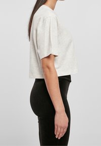 Jasnoszara, krótkotrwała koszulka z krótkim rękawem, wykonana z miękkiego materiału. Połączona z obcisłymi czarnymi spodniami. Widok z boku ukazuje dopasowanie i kształt.