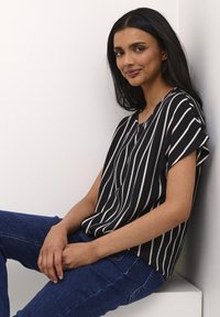 Blusa a righe bianco e nero con maniche corte, scollatura a V e vestibilità rilassata, abbinata a jeans blu scuro, indossata su uno sfondo neutro.