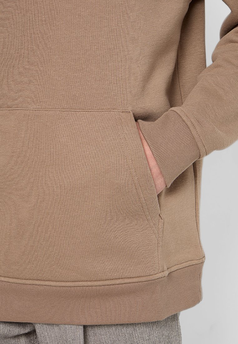 Bruine, gestructureerde pullover met een kangoeroezak en ribgebreide manchetten. De stof is zacht met een subtiel patroon, wat dimensie aan het ontwerp toevoegt.