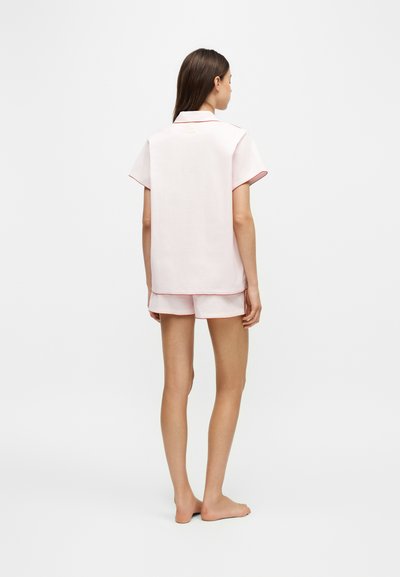 Femme debout pieds nus, de dos, portant un t-shirt rose clair à manches courtes et un short assorti avec un liseré rouge aux manches et à l'ourlet.