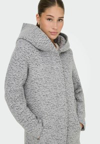 Manteau oversize gris clair en tissu texturé, avec une grande capuche et des poches latérales. Il dispose d'une fermeture à boutons et d'un motif délicatement moucheté.
