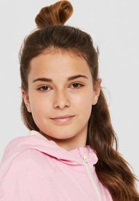 Ljusrosa huvtröja med en vit dragkedja, med en texturerad huva och axelparti, bärs av en person med långt, vågigt brunt hår.