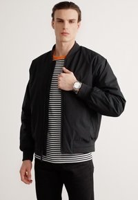 Veste bomber noire en tissu lisse, avec une fermeture éclair sur le devant, des poignets côtelés et une silhouette décontractée, portée sur une chemise rayée.