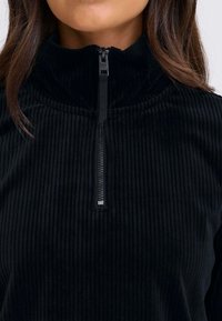 Zwarte corduroy pullover met hoge hals en ritssluiting, voorzien van een textuur met ribbels. Heeft een zilverkleurige metalen rits voor de sluiting en een gladde binnenkant.