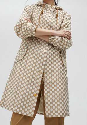 Osoba nosící béžový a bílý kostkovaný kabát s kapucí se stahovacími tkanicemi, zlatými knoflíky a elastickými manžetami, v kombinaci s hnědými kalhotami.