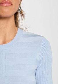 Pull bleu clair à côtes avec un motif à rayures horizontales, doté d'un col rond et d'une texture lisse. Boucle d'oreille sphérique en argent visible.