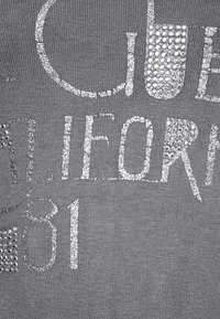 Grått tyg med en slät yta som har djärva, silverfärgade rhinestones som stavar "CALI" och "81", vilket skapar en framträdande grafisk design.