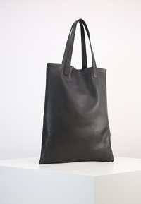Sac fourre-tout en cuir noir texturé avec deux longues poignées, forme rectangulaire et design minimaliste, posé sur une surface blanche.