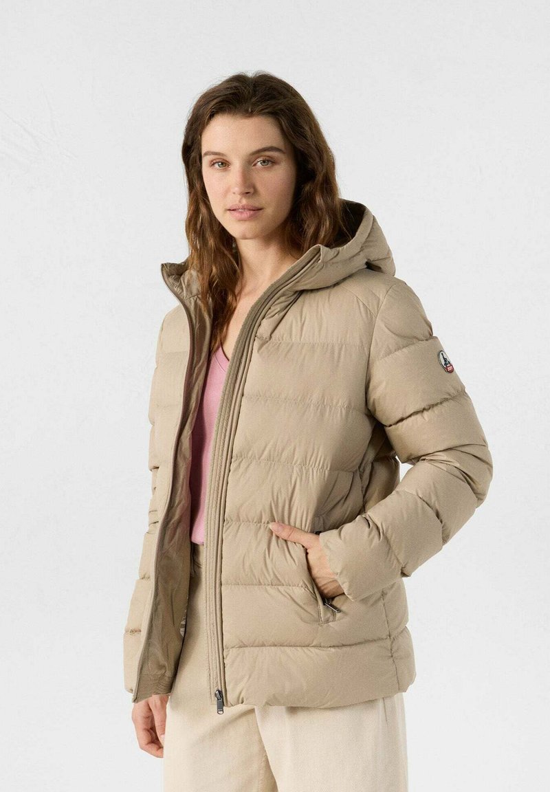Frau trägt eine beige Steppjacke mit Kapuze, ein pinkes Oberteil und hellfarbene Hosen und steht vor einem schlichten weißen Hintergrund.