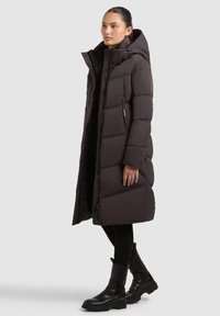 Abrigo largo y acolchado de color marrón oscuro con capucha, con un diseño acolchado, cremallera frontal y cierre a presión. Combinado con botas de tobillo negras.